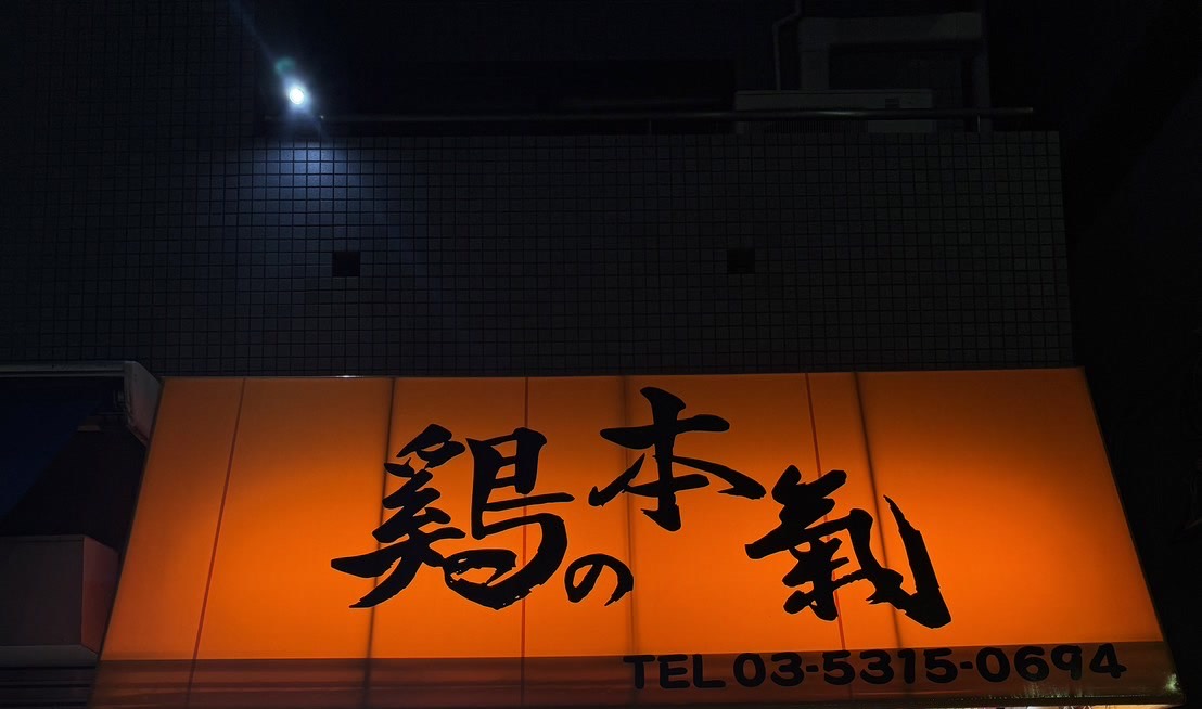 夜の店先。暗いタイル壁の下で橙色のテント看板が光り、黒い筆文字で「鶏の本氣」、下に電話番号。