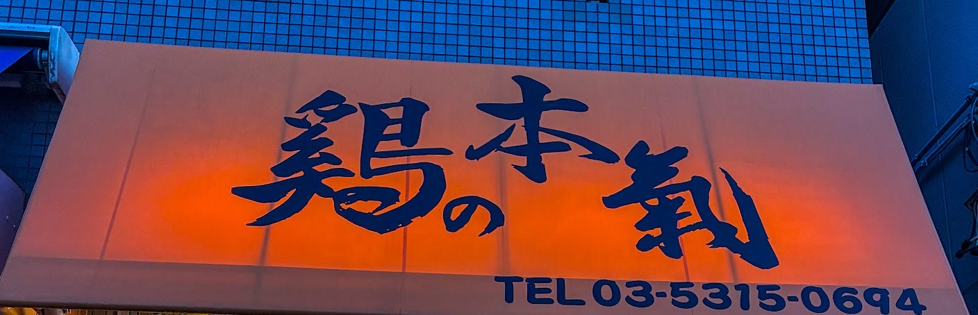 夕暮れの店先。青いタイル壁に橙色のテント看板、濃紺の筆文字で「鶏の本氣」と電話番号。