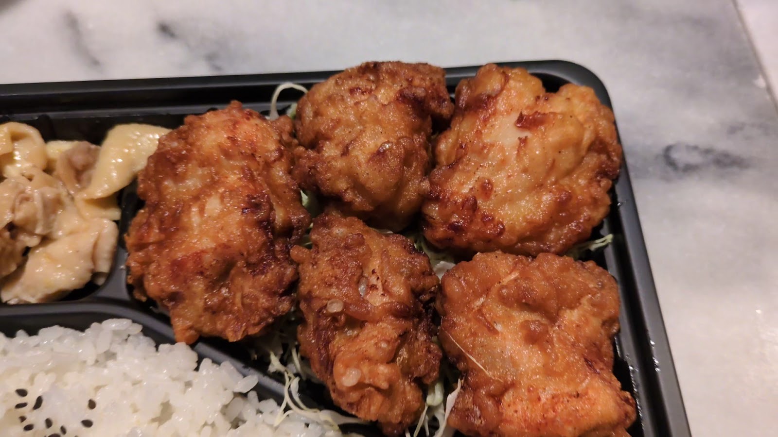 黒い弁当箱に、千切りキャベツの上の唐揚げ5個、黒ごまをふった白いご飯、左に鶏の副菜。